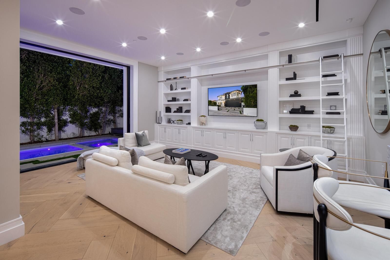 A modern living room with a white sofa, a coffee table, and a TV mounted on the wall. There is a pool visible through the glass wall.