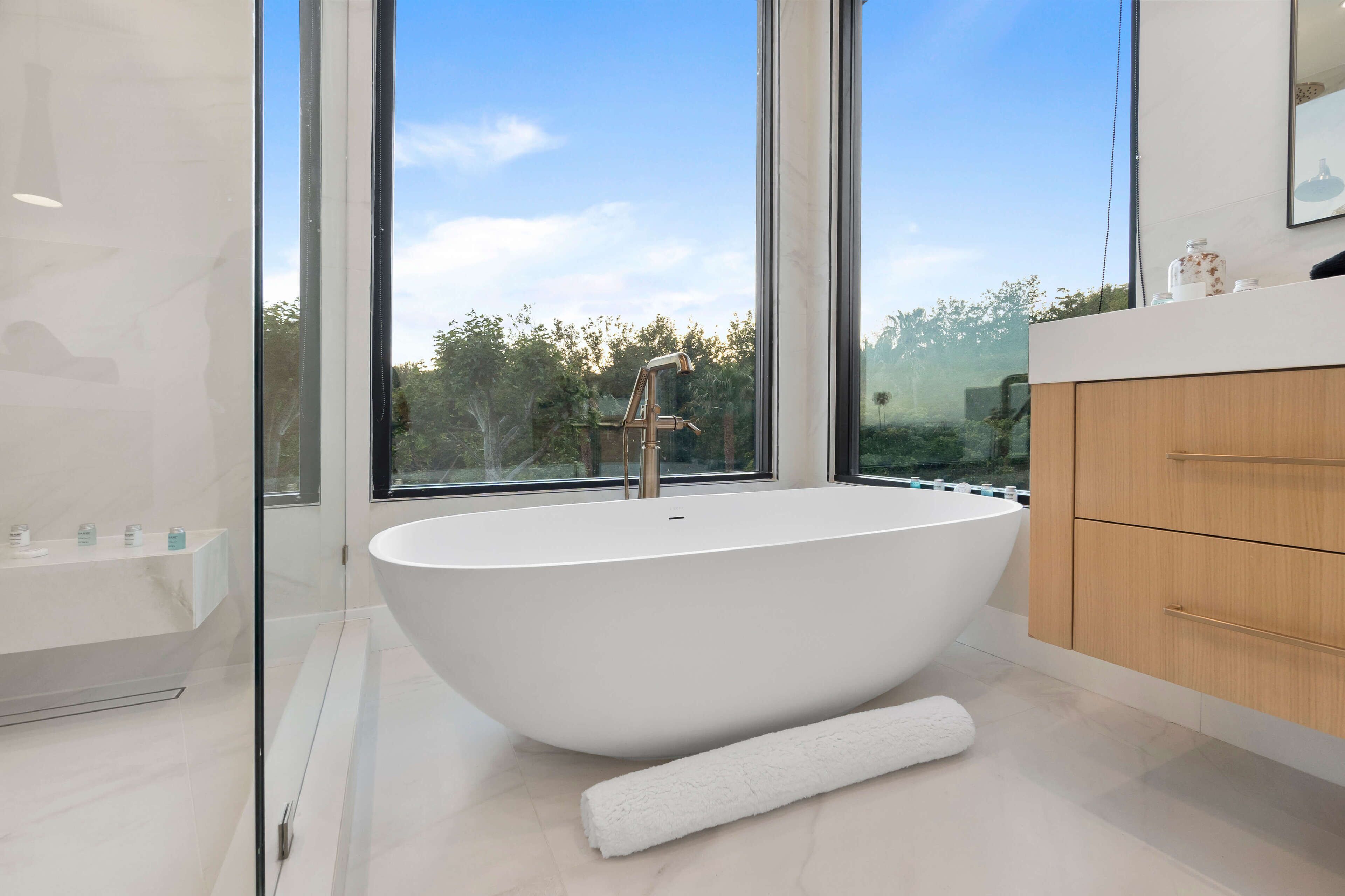 Un baño moderno con una bañera independiente, un box de ducha con mampara de vidrio y un ventanal con vistas a zonas verdes.