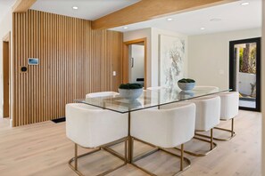 A modern dining area with a glass table, white upholstered chairs, and a wooden accent wall. - Sea Vista (Malibu)