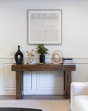 A wooden console table with a framed artwork above it, a vase, a plant, and a stack of books. - Emerson Estate (Sherman Oaks)