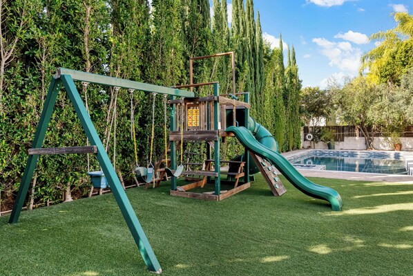 A playground with a swing set and slide, surrounded by a pool and greenery. - Emerson Estate (Sherman Oaks)
