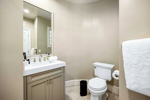 A bathroom with a white toilet, a sink with a mirror above it, and a trash bin. - Emerson Estate (Sherman Oaks)