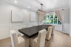 A dining room with a large table and six chairs, white walls, and a view through large windows. - Emerson Estate (Sherman Oaks)