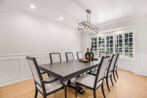 A dining room with a dark wooden table and six chairs, white paneled walls, and a large window with a view of greenery. - Emerson Estate (Sherman Oaks)