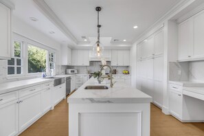 Una cocina amplia con muebles blancos, una isla de mármol y una lámpara colgante.