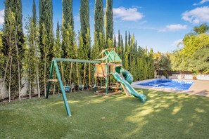 Un parque infantil con tobogán, columpio y estructura para escalar, rodeado de árboles altos y una piscina.
