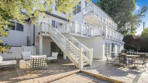 Een wit, twee verdiepingen tellend gebouw met een trap die naar een balkon leidt, een buiten zitgedeelte met tafels en stoelen, en een overdekt terras.