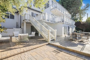 Un edificio blanco de dos pisos con una escalera que conduce a un balcón, zona de estar exterior con mesas y sillas, y un porche cubierto.