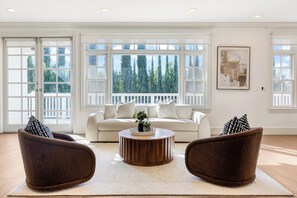 Un salón amplio con un sofá blanco, dos sillones marrones, una mesa de centro de madera y ventanas grandes que ofrecen vistas a árboles.
