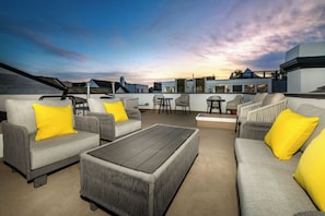 An outdoor seating area with grey sofas and yellow cushions, a wooden coffee table, and a view of buildings in the background. - Reef Mall (Marina del Rey)