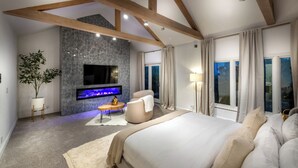 Un dormitorio moderno con una cama grande, un televisor de pantalla plana montado en una pared con textura, una zona de estar con una mesa redonda y vistas al exterior a través de amplios ventanales.