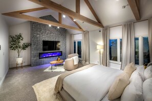 A modern bedroom with a large bed, a flat-screen TV mounted on a textured wall, a seating area with a round table, and a view of the outdoors through large windows. - Reef Mall (Marina del Rey)
