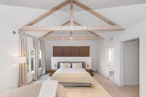 A bedroom with a high ceiling, exposed wooden beams, a large bed with a headboard, bedside tables, and a window with curtains. - Reef Mall (Marina del Rey)