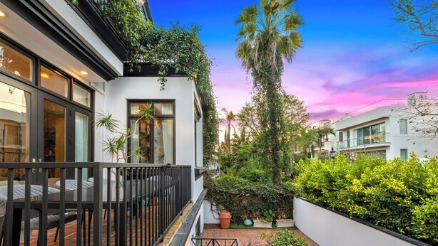 Un edificio moderno con grandes ventanales, un balcón y un jardín con diversas plantas y una palmera alta.