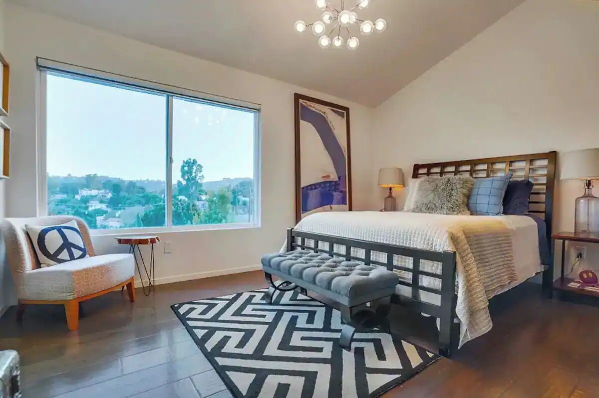 Un dormitorio con un ventanal grande, una cama con cabecera, un banco, una alfombra y una araña.