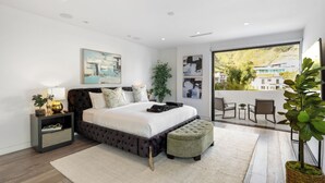 A modern bedroom with a large bed, a nightstand, a bedside table, a window with a view of buildings, and a balcony with chairs. - Franklin Modern (West Hollywood)