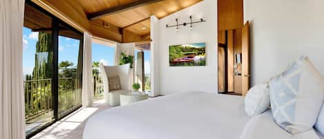 A bedroom with a large bed, a view of the outdoors, a small seating area, and a painting on the wall.