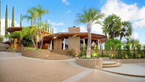 Una casa moderna con fachada de piedra, una terraza de madera y un jardín ajardinado con palmeras.