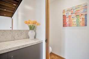 A bathroom with a marble countertop, a white vase with flowers, and a colorful painting on the wall. - Via Vallarta (Encino)