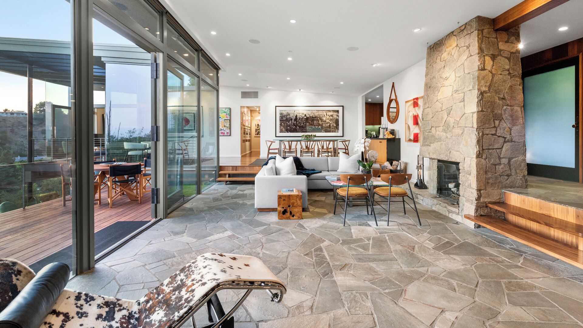 Uma sala de estar moderna com lareira de pedra, porta de vidro para um deck e área de jantar com móveis de madeira.