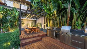 An outdoor kitchen area with a grill, wooden deck, and lush tropical plants.