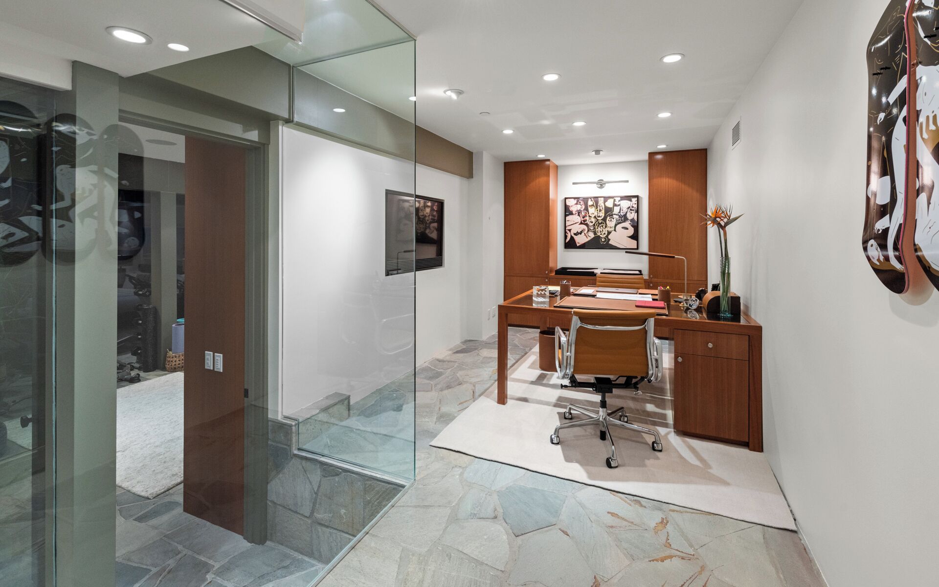 A modern office space with a glass partition, a desk with a chair, a wooden cabinet, and a wall-mounted artwork.