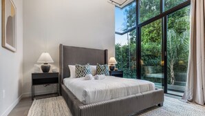 Quarto moderno com uma cama grande, duas mesas de cabeceira com abajures, um tapete estampado e vista para o verde através de amplas janelas.