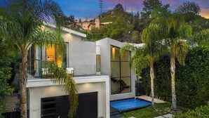 Uma casa moderna de dois andares com piscina, rodeada por vegetação exuberante e palmeiras.