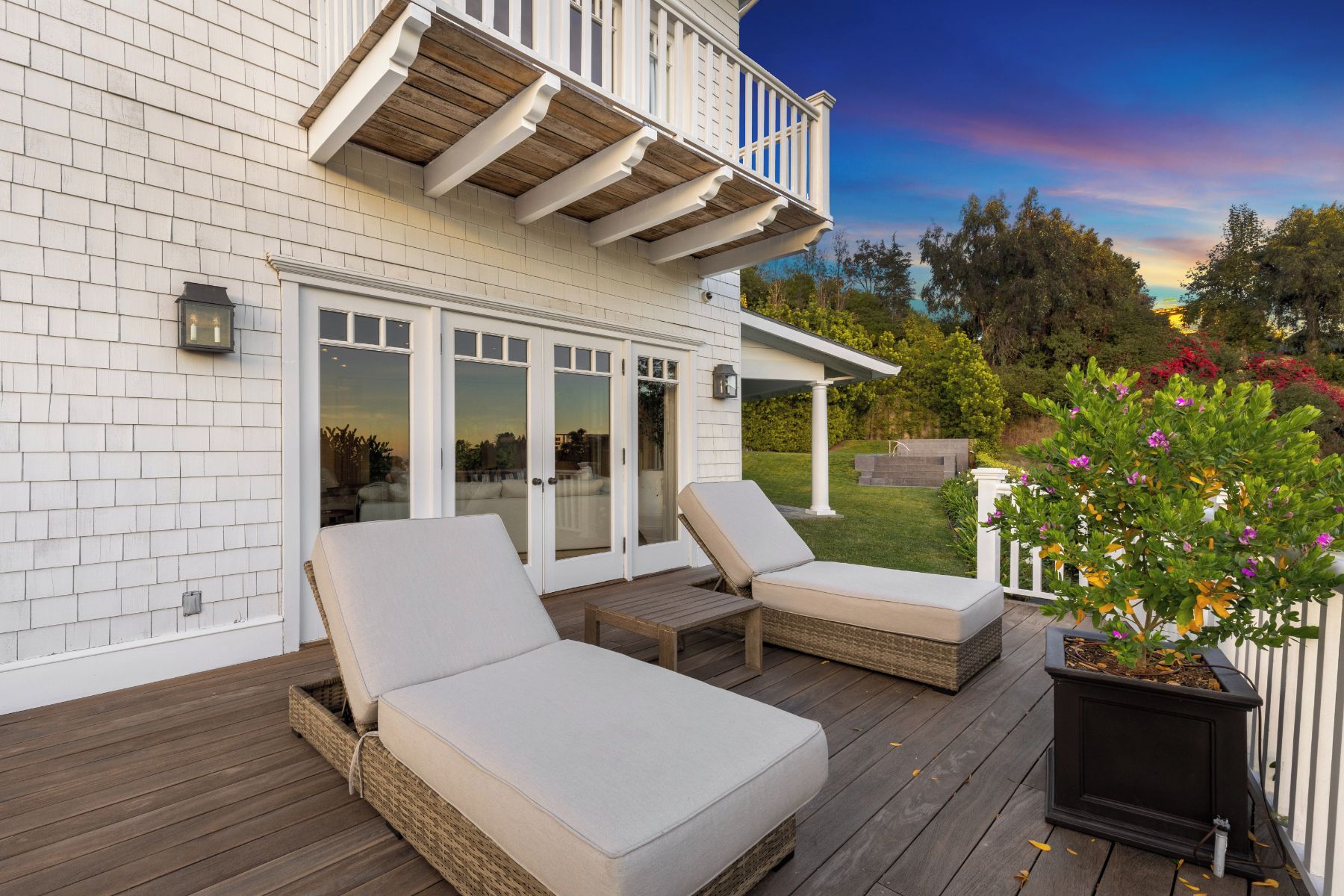 A deck with two lounge chairs and a small table, a white house with a balcony, and a garden with various plants and flowers.