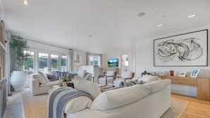 Un salon spacieux avec un grand canapé blanc, une table basse et une vue sur l’extérieur grâce à de grandes fenêtres.