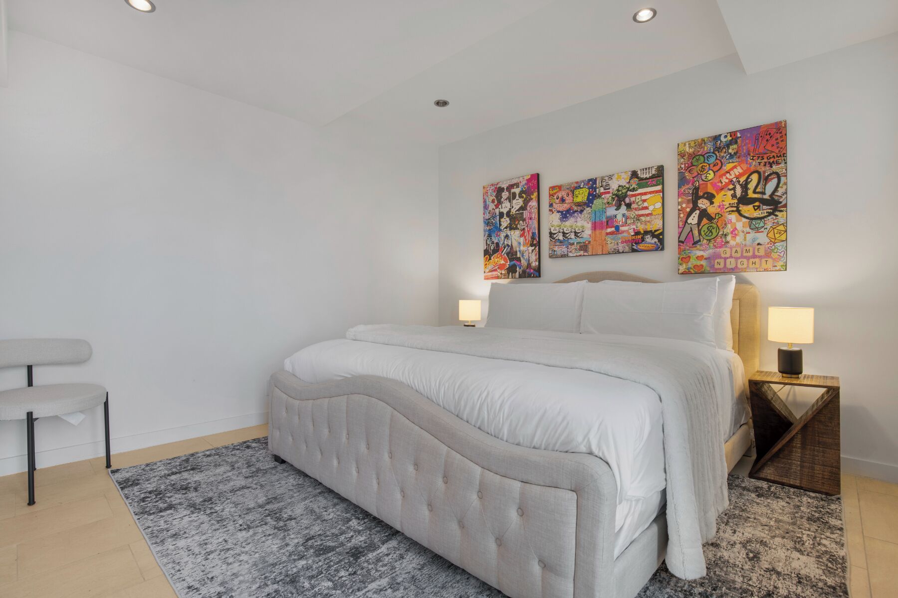A bedroom with a large bed, two bedside lamps, a grey upholstered bench, and three colorful posters on the wall.