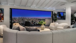 Uma sala de estar moderna com um sofá grande, uma mesa de centro e vista para o horizonte urbano ao anoitecer.