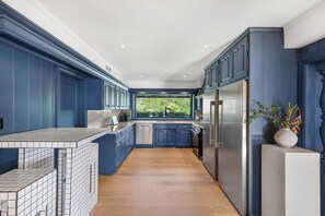 A modern kitchen with blue cabinetry, a stainless steel refrigerator, and a large window offering a view of greenery. - Beverly Tree House (Beverly Hills)