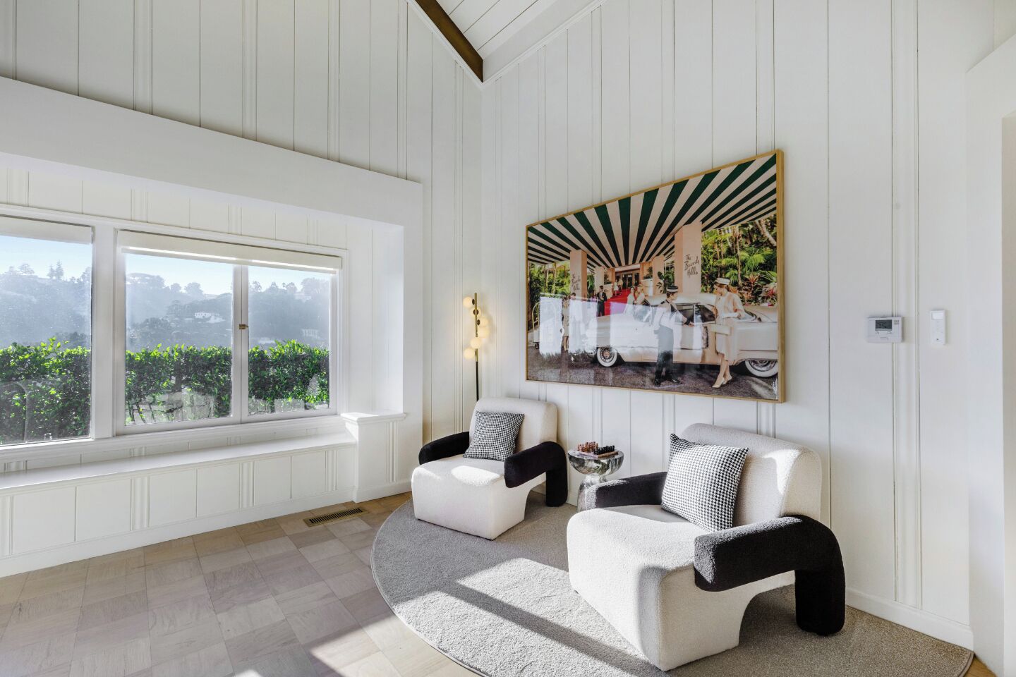 A room with white paneled walls, a large window with a view of greenery, two white armchairs with black and white cushions, and a large framed picture on the wall.