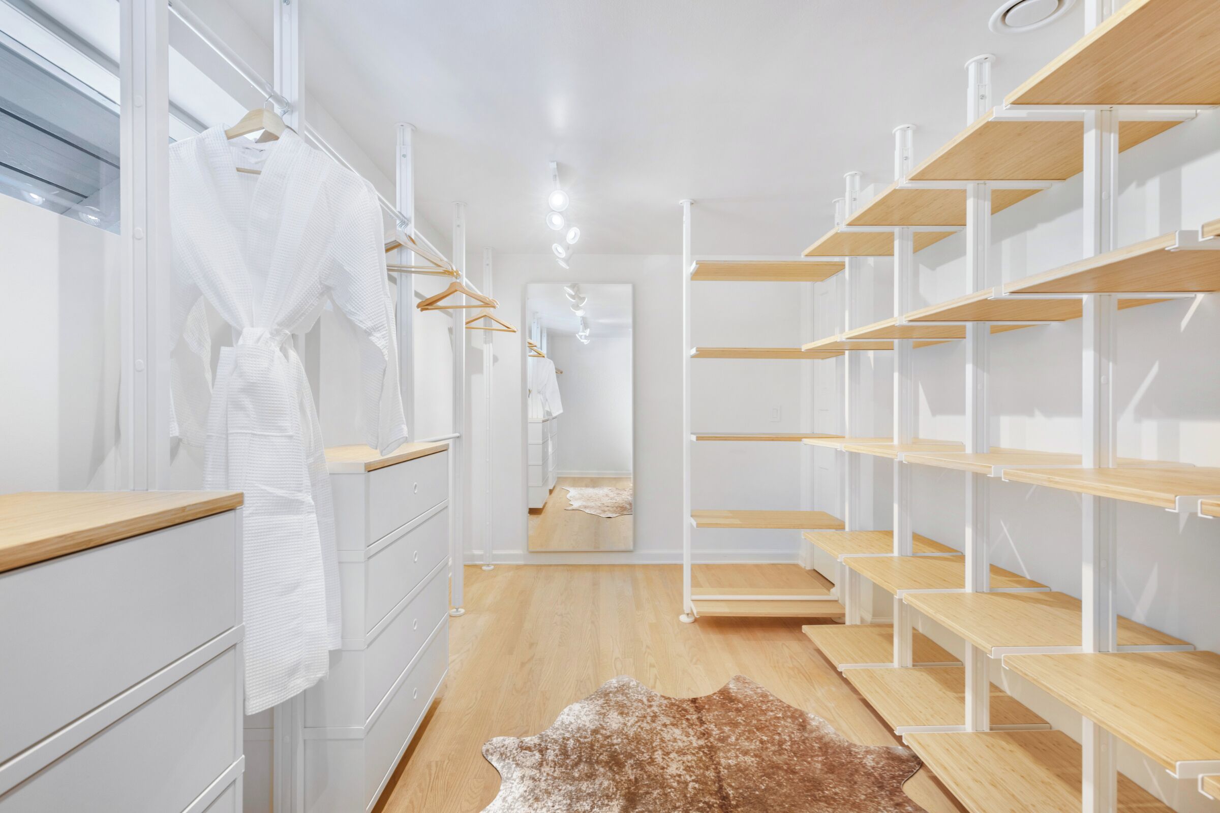 A modern, well-lit walk-in closet with white cabinetry, wooden shelves, and a leather rug on the floor.