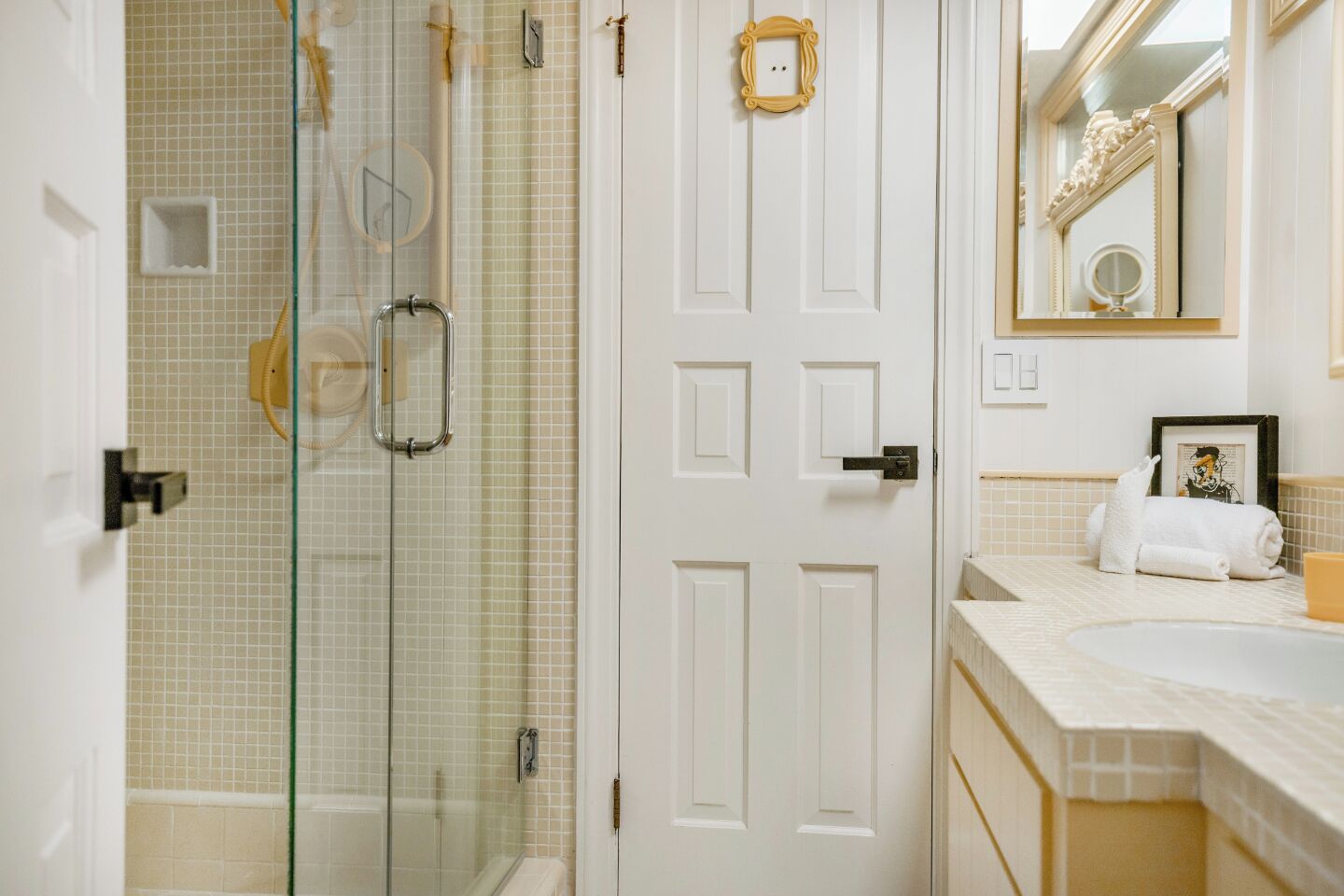 Un baño con cabina de ducha de vidrio, una puerta blanca con marco dorado, un lavabo con espejo y una toalla sobre el encimera.