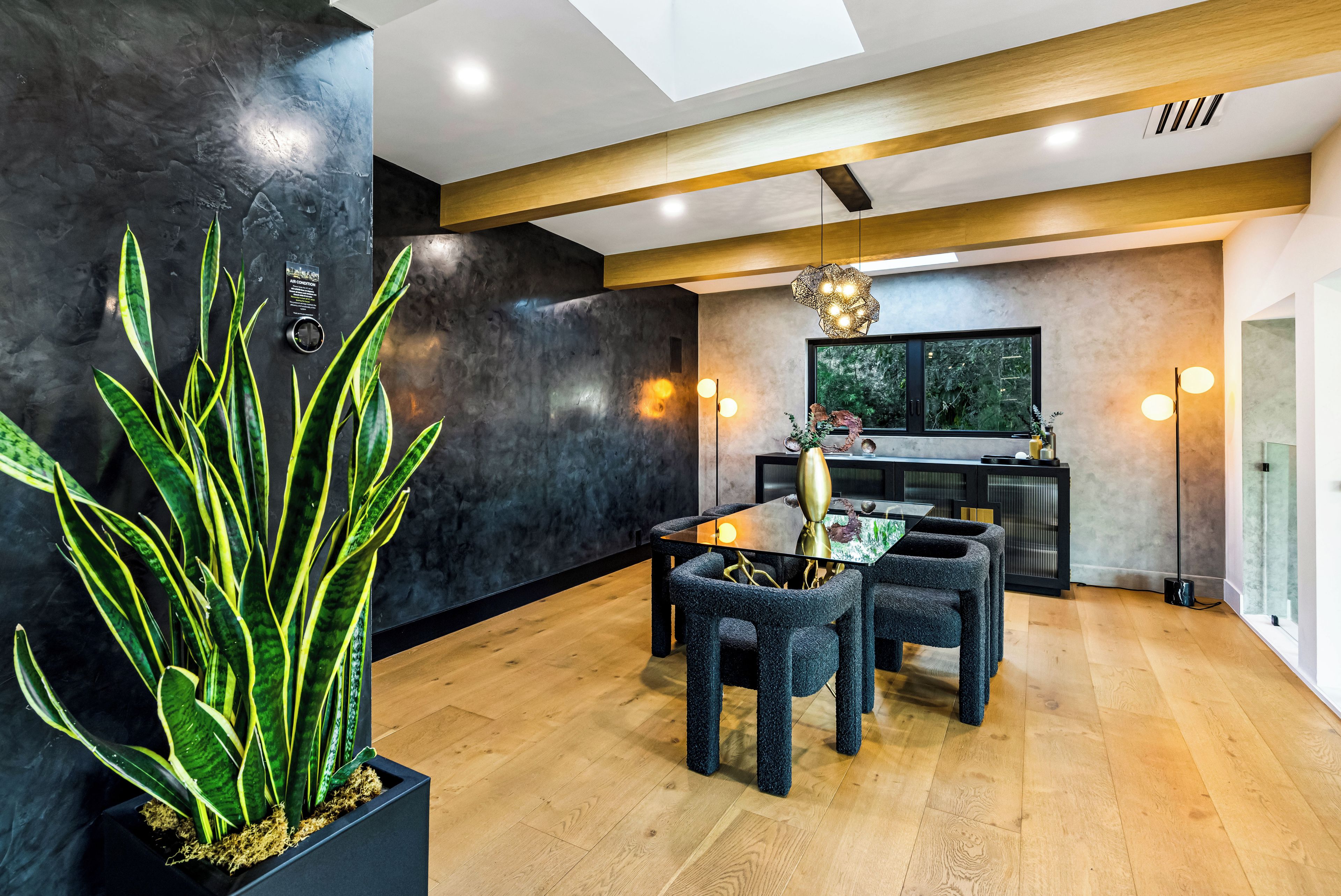 Un espace repas moderne avec un mur noir, une grande plante en pot, une table à plateau en verre et des chaises sombres.