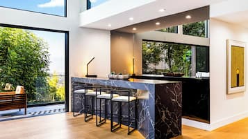 A modern kitchen with a marble island, bar stools, and a view of greenery outside.