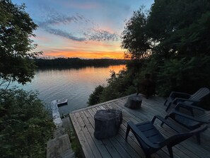 Une terrasse en bois avec des chaises et un foyer, donnant sur un lac paisible au coucher du soleil.