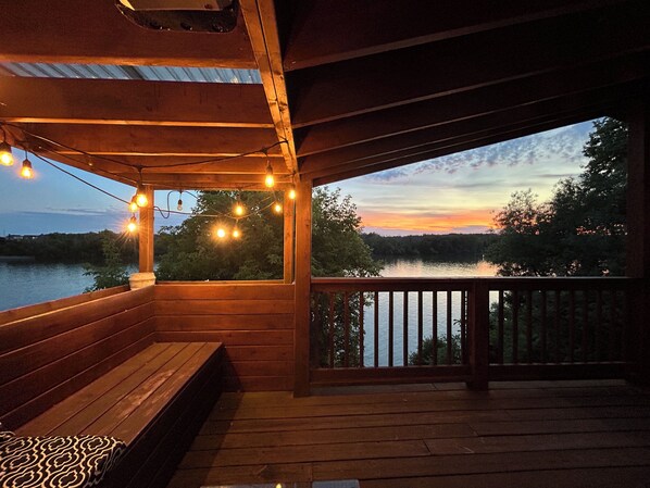 A wooden deck with a bench, string lights, and a view of a lake at dusk.