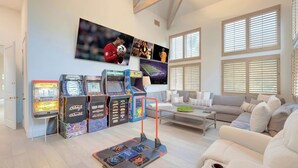 A living room with a white couch, arcade machines, and a large TV on the wall. - Carolwood Estate (Beverly Hills)
