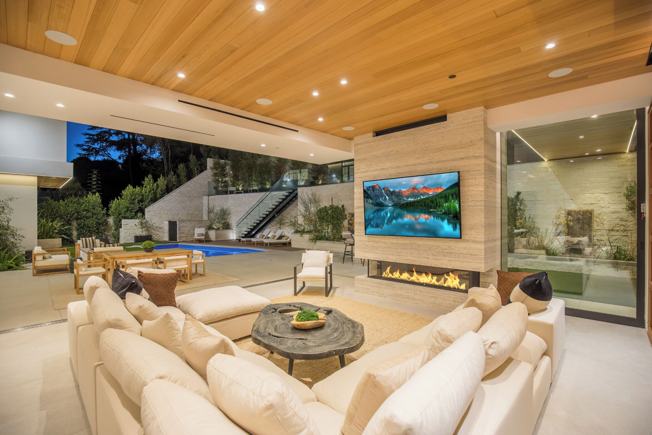 A modern living room with a fireplace, a large TV, and a pool area visible through the glass walls.