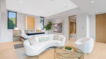 A modern living room with a white sofa, a glass coffee table, and a bed with a blue headboard.