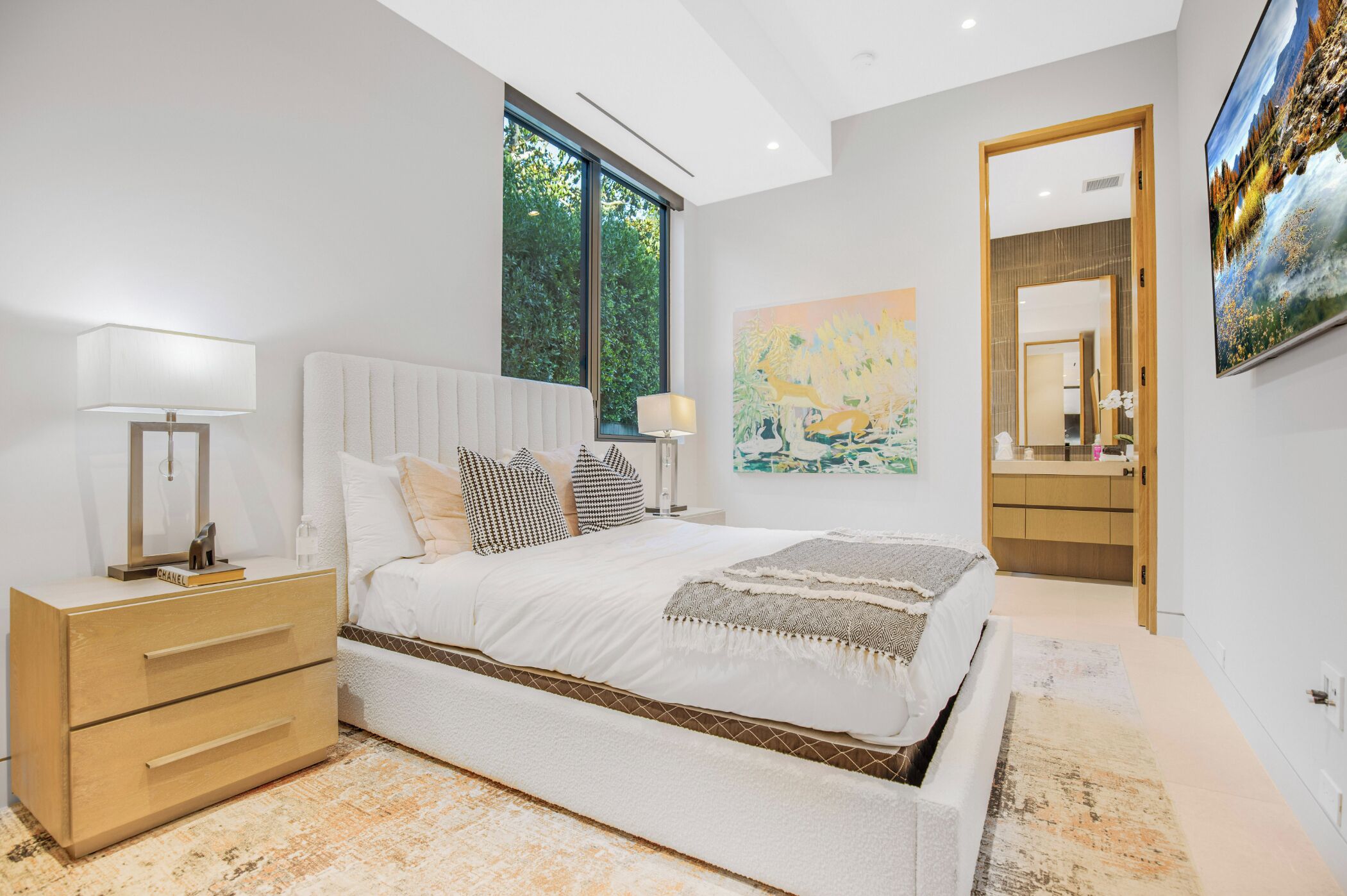 Un dormitorio moderno con una cama grande, mesitas de noche, una lámpara y una ventana con vistas a árboles, además de un cuadro en la pared.