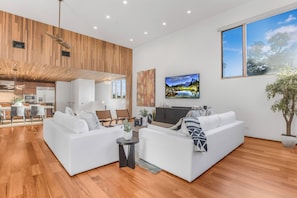 A modern living room with a large white sectional sofa, a flat-screen TV mounted on the wall, and a dining area with a wooden ceiling. - Cedar Loft Villa (Venice)