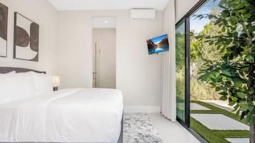 A modern bedroom with a large bed, a flat-screen TV mounted on the wall, and a view of a landscaped garden through a sliding glass door.