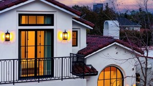 A white building with a red-tiled roof, two lit windows, and a balcony with a black railing. - Villa Camellia (Studio City)