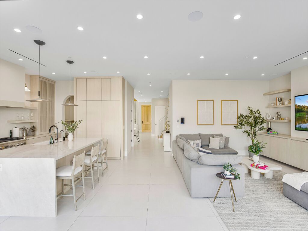 A modern kitchen with a central island, a dining area with chairs, a living room with a sofa and a coffee table, and a TV on the wall.