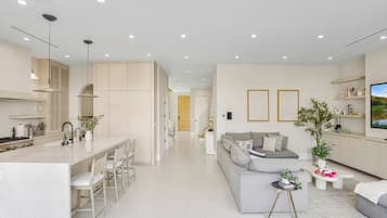 A modern kitchen with a central island, a dining area with chairs, a living room with a sofa and a coffee table, and a TV on the wall.