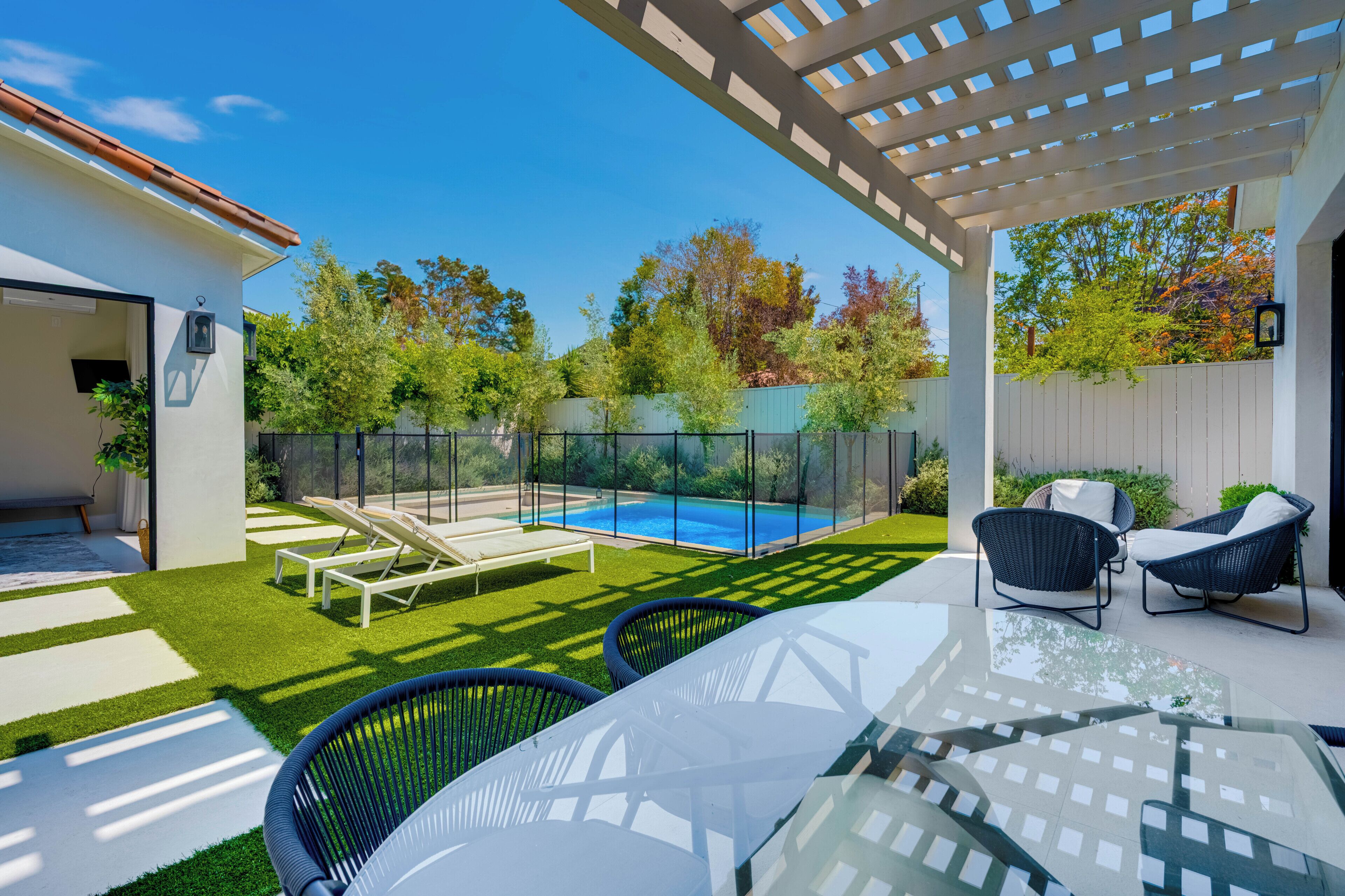 A modern outdoor patio with a glass table, two wicker chairs, a pool, and a pergola.
